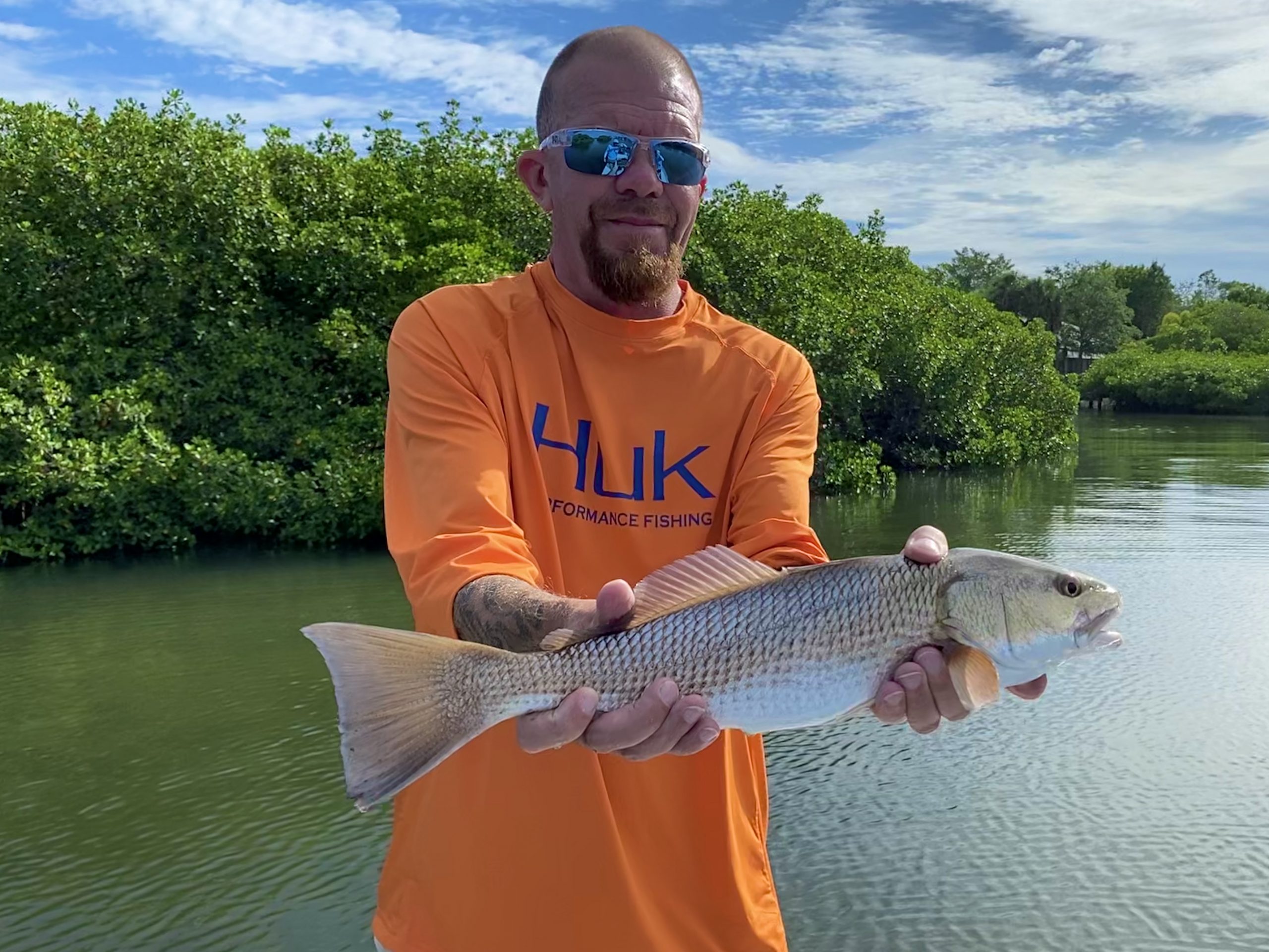 A bald angler wearing oakley sunglasses and a bright orange shirt holds a redfish for the camera