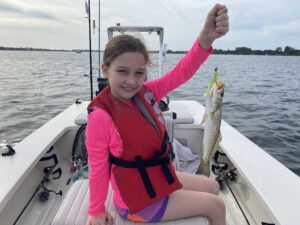 A young girl sits in a boat and holds up a spotted seatrout for the camera that she caught