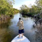 An angler fishes from the bow of a shallow water poling skiff in a narrow mangrove passage