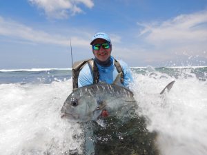 An angler holds a grand trevally up for the camera in the waves of the seychelles